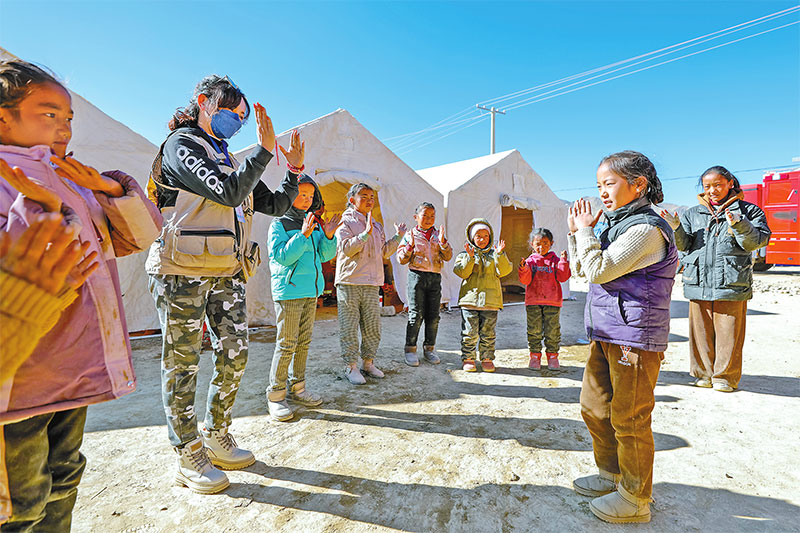 འདི་ནི་དིང་རི་རྫོང་ཆུ་ལྷོ་ཤང་མཚོ་སྔ་གྲོང་ཚོའི་གནོད་འཚེ་ཕོག་པའི་མང་ཚོགས་བཀོད་སྒྲིག་བྱ་ཡུལ་དུ་ཐན་ཞི་རེན(གཡོན་གྱི་གཉིས་པ)གྱིས་ཕྲུ་གུ་ཚོ་དང་མཉམ་དུ་ཞབས་བྲོ་འཁྲབ་བཞིན་པ།